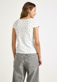 Hvid bomulds T-shirt med marineblå blomstermotiv, korte ærmer, rund halsudskæring; parret med højtaljede grå denimbukser med baglommer.