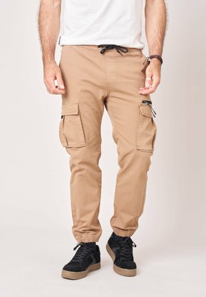 Man die beige cargobroek met trekkoord in de taille en zwarte sneakers draagt, staande tegen een effen lichte achtergrond.