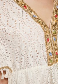 Witte kanten top met bloemmotieven en een gouden geborduurde halslijn met rode, blauwe en groene accenten; geplooide zoom met franjes.