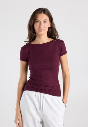 Jonge vrouw met lang bruin haar, gekleed in een aansluitend bordeauxrood shirt met korte mouwen en lichtgrijze joggingbroek, staand met handen in de zakken.