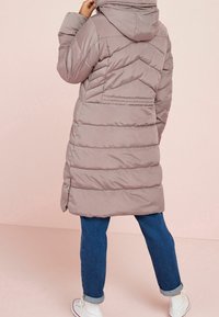 Person wearing a mauve puffer coat with hood, blue rolled-up jeans, and white sneakers, standing against a pale pink background.
