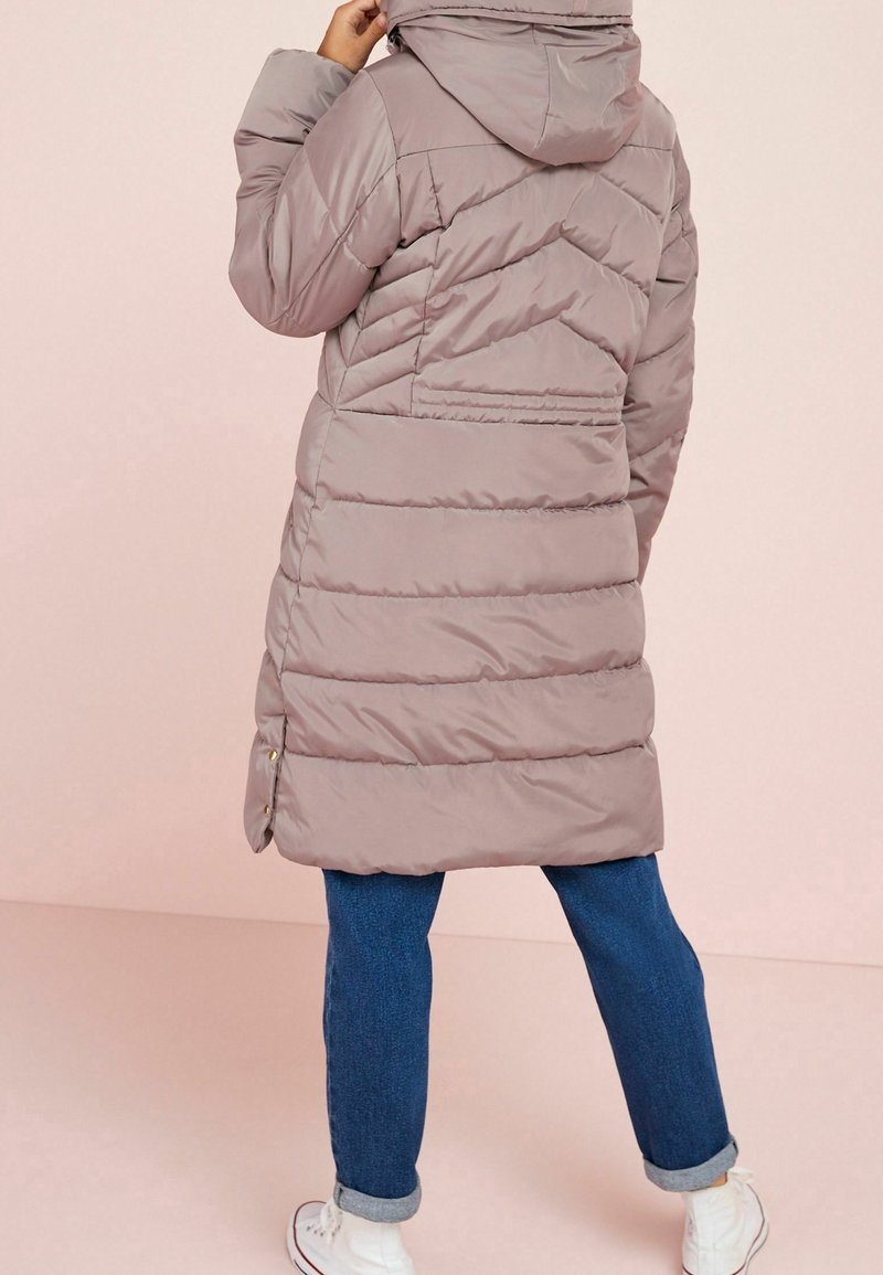 Person wearing a mauve puffer coat with hood, blue rolled-up jeans, and white sneakers, standing against a pale pink background.