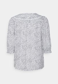 Blusa branca com bolinhas pretas, apresentando uma parte de trás franzida, mangas três quartos e fecho de botão no pescoço. Textura de tecido suave.