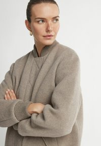 Beżowa, wełniana kurtka bombowca w oversize'owym kroju, z prążkowanym kołnierzem i mankietami, z pikowanym przednim panelem. Miękka tekstura, luźny krój.