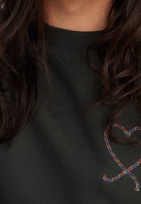 Černá mikina s kulatým výstřihem, zdobená mnohobarevným vyšitým srdcem na hrudi. Hladká textura, ležérní vzhled.