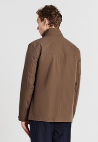 Giacca lunga marrone, con maniche lunghe e colletto alto, realizzata in tessuto liscio. Presenta cuciture in rilievo e un taglio dritto. La vista posteriore mostra dettagli minimi.