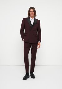 Burgundy double-breasted suit jacket with black satin lapels, paired with matching tailored pants and black loafers. Light pink shirt underneath.