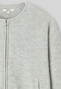 Veste en laine grise avec un col arrondi, fermeture éclair à l'avant et poches latérales. Texture douce avec un design minimaliste.