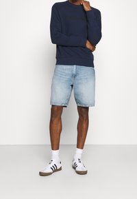 Námořnická mikina s kulatým výstřihem a embosovaným logem, spárovaná s lehkými denimovými šortkami. Bílé tenisky s tmavými pruhy a bílé ponožky dotvářejí vzhled.