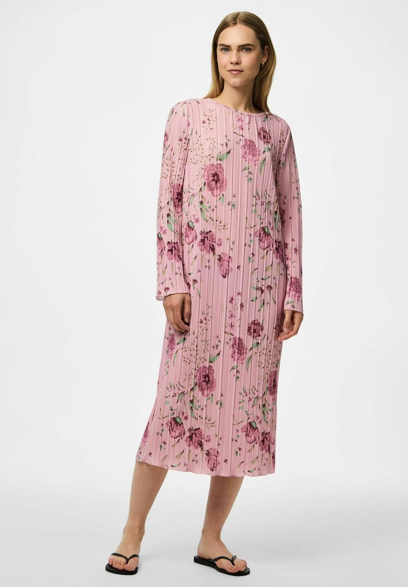 Långärmad klänning i ljusrosa med veckat mönster, prydd med blommor i nyanser av lila och grönt. Bärs med svarta flip-flops.