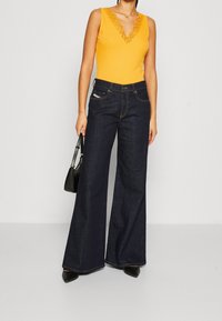 Gelbes ärmelloses Top mit Spitzenbesatz, kombiniert mit dunkelblauen weiten Jeans. Das Model hält eine kleine schwarze Handtasche und trägt schwarze spitze Schuhe.