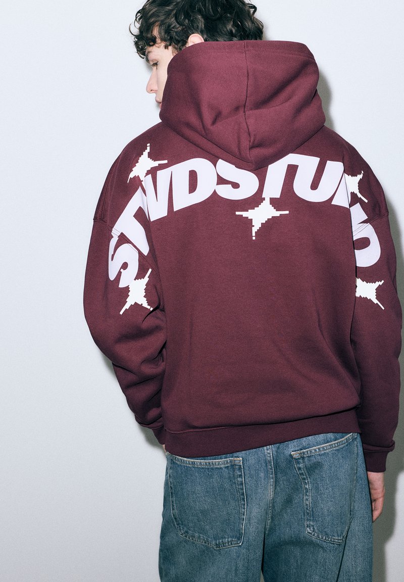 Bordeauxrode capuchontrui met witte grafische tekst en sterpatronen op de achterkant. Heeft lange mouwen en geribbelde manchetten. Draagt bij een spijkerbroek.