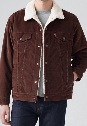 Homme portant une veste en velours côtelé marron avec un col en polaire blanche par-dessus une chemise grise et un jean bleu foncé, main dans la poche d'un côté.