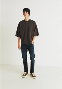 Barna oversized póló, sötét farmernadrág és barna sportcipő fehér cipőfűzővel. Egyszerű, díszítetlen dizájn, könnyű anyag, laza szabás.