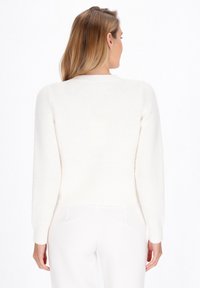 Vrouw met schouderlang blond haar draagt een witte gebreide trui en witte broek, staand met haar rug naar de camera tegen een witte achtergrond.