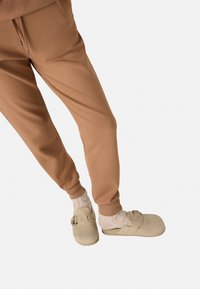 Pantalones de chándal marrones combinados con zuecos beige. Los zuecos tienen una correa y un detalle de hebilla. Se llevan calcetines de color claro y texturizados.