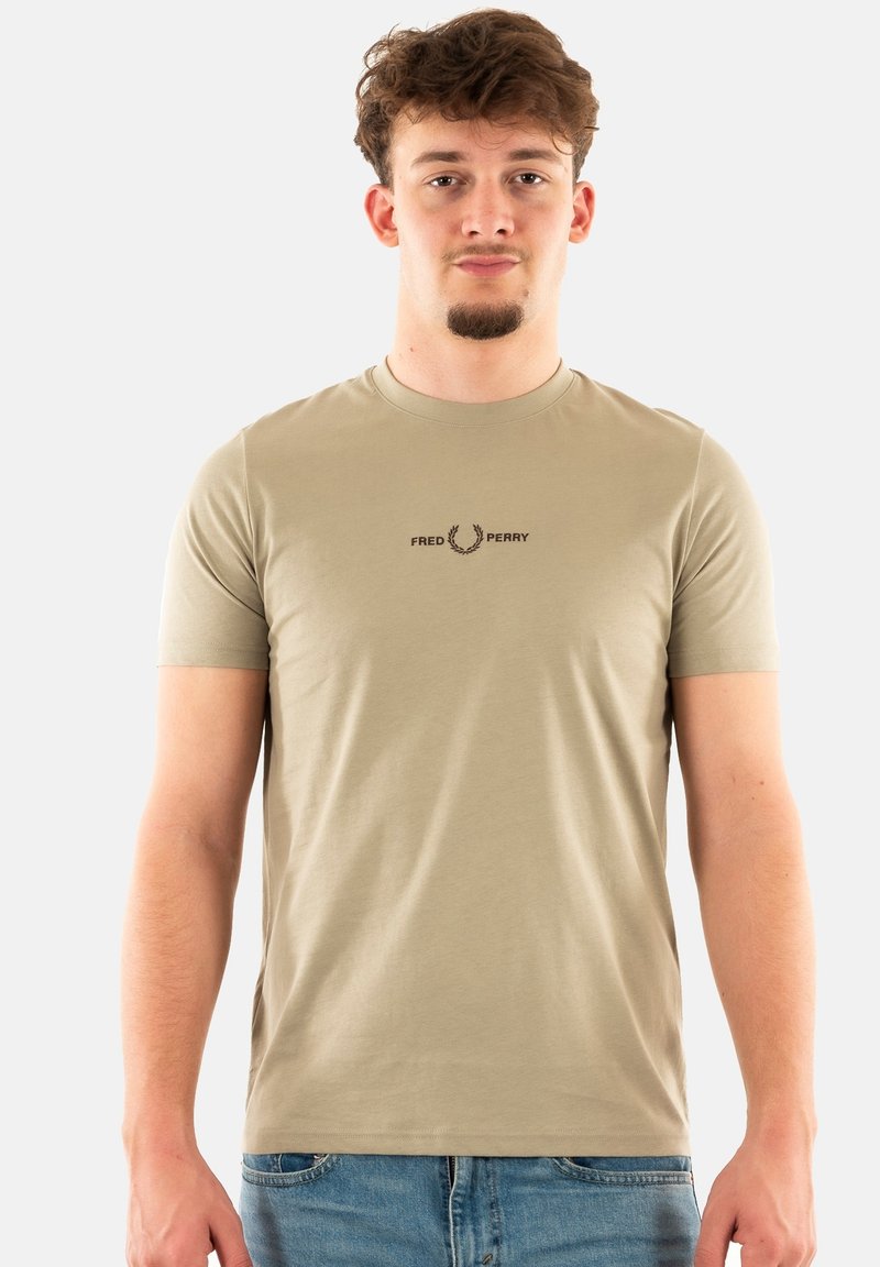 Camiseta de algodón de color beige claro con cuello redondo, mangas cortas y un pequeño logo marrón centrado en el pecho. Textura suave.