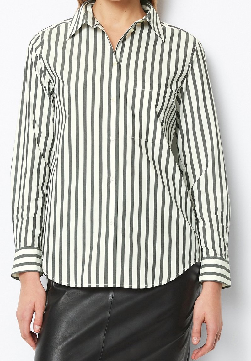 Femme portant une chemise à manches longues boutonnée avec des rayures verticales noires et blanches et une jupe en cuir noire.
