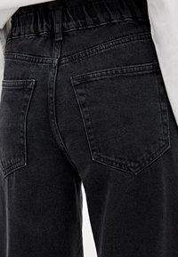 Pantalon en jean noir avec une ceinture extensible, deux poches arrière de forme rectangulaire, et une finition texturée subtile.