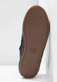 Black suede slip-on shoe with woven design, featuring a rubber outsole. UGG logo embossed on tan sole with textured pattern.