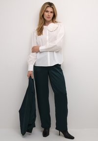 Hvid skjorte med knap ned foran og en tekstureret blomsterdetalje, parret med mørkegrønne bukser med vidde og sorte højhælede støvler, mens der holdes en matchende blazer.