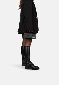 Botas negras de cuero hasta la rodilla con una textura suave y suelas gruesas, combinadas con un bolso estructurado negro con detalles dorados.