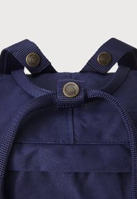 Mochila navy escura com alças trançadas, detalhes em metal latão e uma textura de tecido suave. Apresenta um fecho de cordão e costura reforçada.