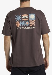 Camiseta de algodón marrón con un diseño gráfico colorido que presenta formas abstractas y el texto "BILLABONG" impreso en la parte posterior.