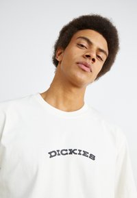 White cotton t-shirt featuring a curved black "DICKIES" logo on the front. The design includes a round neckline and short sleeves.
