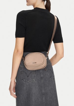 Frau trägt ein schwarzes Kurzarmoberteil und einen grauen Jeansrock mit einer beigefarbenen Umhängetasche, die am unteren Rücken positioniert ist.