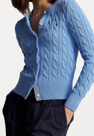 Frau trägt eine hellblaue, kabelgestrickte Strickjacke mit Knöpfen, eine Hand in der Tasche, kombiniert mit dunklen, plissierten Hosen vor einem schlichten Hintergrund.