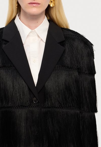 Zwarte fringed jacket met gestructureerde kraag en knoopsluiting, gelaagd ontwerp, gedragen over een witte overhemd met kraag. Geverfd met zwarte franjes.
