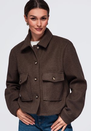 Femme portant une veste en laine marron foncé boutonnée avec deux poches à rabat devant et un jean bleu, debout, les mains sur les hanches.