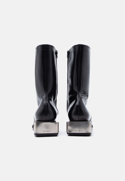 Μαύρες μπότες μήκους μέχρι τη μέση της γάμπας με γυαλιστερά μεταλλικά χοντρά τακούνια και φερμουάρ στο πίσω μέρος, εμφανίζονται από πίσω σε ένα απλό λευκό φόντο.