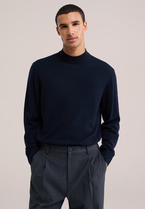 Homme aux cheveux courts et foncés portant un pull bleu marine et un pantalon plissé gris foncé, debout les mains dans les poches devant un fond clair.