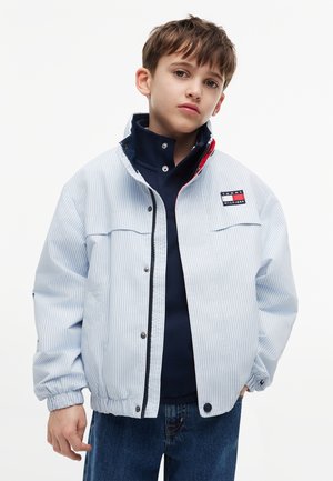 Junge trägt eine hellblaue gestreifte Jacke über einem dunkelblauen Hemd und blauen Jeans, schaut mit neutralem Gesichtsausdruck geradeaus vor weißem Hintergrund.
