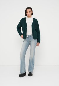 Σκούρο πράσινο υφασμάτινο ζακέτα με δύο κουμπιά, φορεμένο πάνω από λευκή μπλούζα και συνδυασμένο με ανοιχτό μπλε φαρδιά τζιν και μαύρα χοντρά παπούτσια.