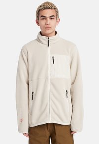 Lysebeige fleece jakke med ståkrave, lynlåslukning foran, to sidelommer og en brystlomme i et kontrasterende crememateriale.