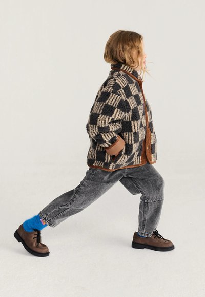 Enfant portant une veste polaire à carreaux, un jean gris, des chaussettes bleu vif et des chaussures marron, marchant vers l’avant sur un fond clair.