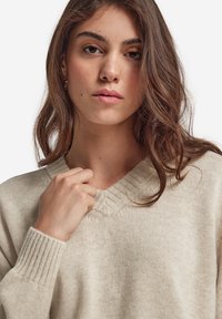 Beige virkade tröja med V-ringning, ribbade manschetter och ett strukturerat mönster. Slät tyg och avslappnad passform, som framhäver kragdetaljen.