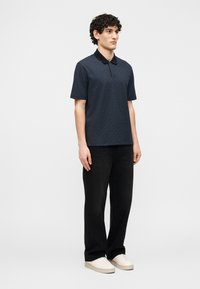 Νεαρός άνδρας στέκεται φορώντας σκούρο πόλο με σχέδια, μαύρο παντελόνι με φαρδιά μπάτζα και λευκά αθλητικά παπούτσια, μπροστά σε απλό λευκό φόντο.