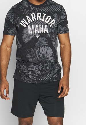 Homme portant un t-shirt noir avec un texte blanc "WARRIOR MANA" et des motifs de feuilles, associé à un short noir uni, debout devant un fond blanc.
