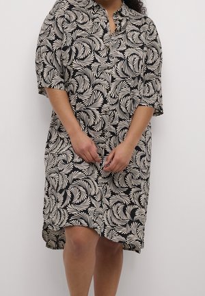 Kobieta w czarno-beżowej sukience zapinanej na guziki z wijącym się wzorem liści, stojąca na tle jednolitego jasnego tła.