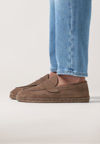 Espadrillas in suede marrone con suola piatta e falda decorativa sulla punta, caratterizzate da rifinitura in juta intrecciata. Indossate con jeans di denim azzurro chiaro.