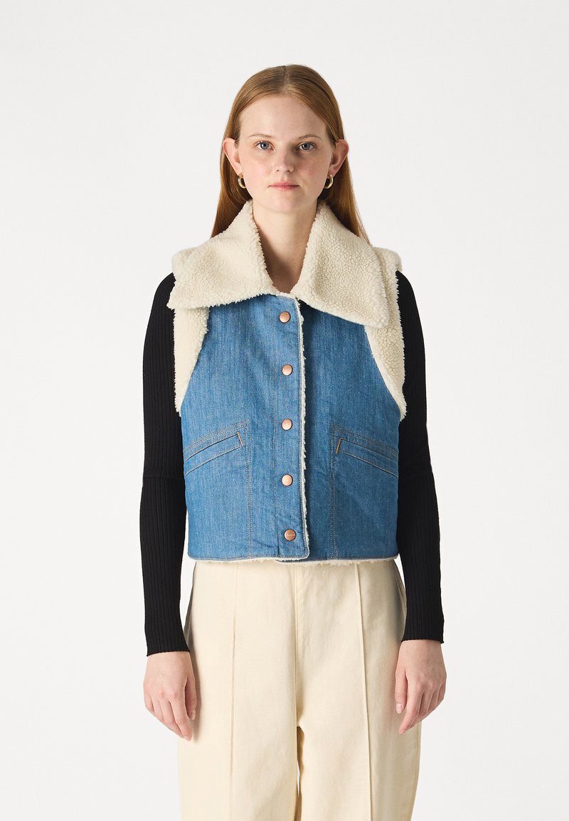 Niebieska dżinsowa kamizelka z kołnierzykiem z kremowego sztucznego baranka, z przodu zapinana na napy i z dwoma bocznymi kieszeniami. Noszona na czarnym, długim topie.