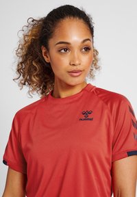 Rotes Sporttop mit Rundhalsausschnitt, kurzen Ärmeln und einer subtilen Textur. Es hat ein marineblaues Logo und Akzente entlang der Ärmel.
