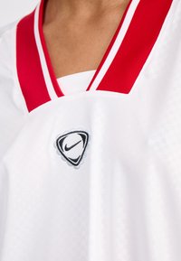 Bílý sportovní dres s červeným límcem ve tvaru V a bílými pruhy. Obsahuje černé a šedé vyšívané logo Nike na oblasti hrudi.