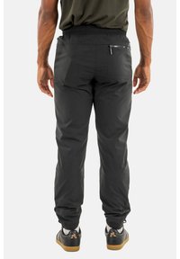 Schwarze elastische Hosen mit Gummizugbund, Reißverschluss auf der Rückseite und schmal zulaufenden Bündchen aus einem glatten, leichten Material. Kombiniert mit lässigen schwarzen Schuhen.