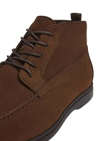 Brązowe zamszowe botki na kostkę o gładkiej fakturze, z wiązaniem i kontrastującą czarną gumową podeszwą. Posiadają pętelkę na pięcie.