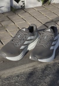 Graue Sportschuhe mit einem texturierten Netz-Obermaterial, weißen Akzentstreifen, abgerundetem Zehenbereich und einer gepolsterten weißen Sohle.